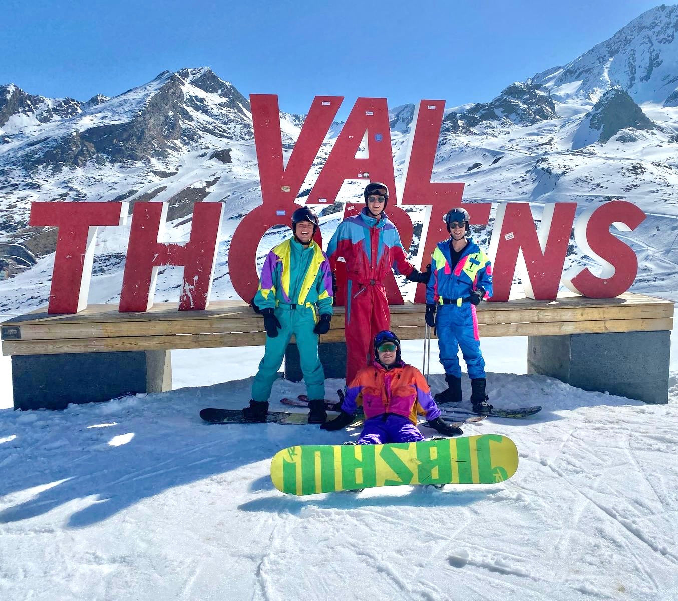Groep jongeren in foute vintage retro skipakken poserend bij Val Thorens bord in de Franse Alpen met snowboard – opvallende wintersport outfits 2025